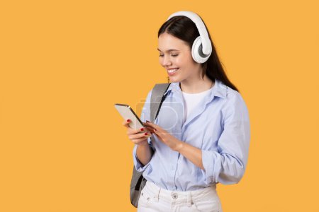 Mujer joven con auriculares absorbidos en el uso de su teléfono inteligente, conectado, viendo podcast sobre fondo amarillo
