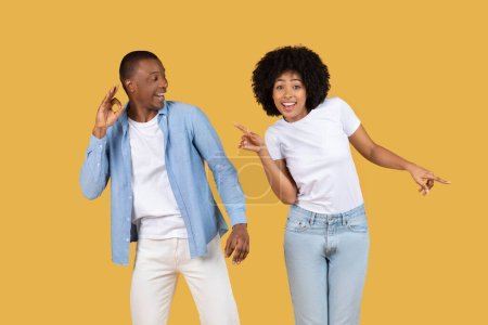 Téléchargez les photos : Un homme et une femme afro-américains souriants vêtus de façon décontractée, dansant et pointant du doigt, profitant sur fond jaune - en image libre de droit