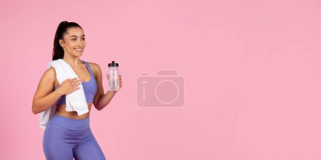 Photo for A smiling fit woman in activewear with a towel and water bottle taking a break on a pink background - Royalty Free Image