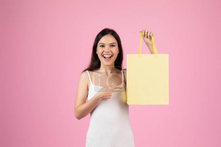 Eine fröhliche junge Frau im weißen Kleid deutet auf eine leuchtend gelbe Einkaufstasche vor rosa Hintergrund