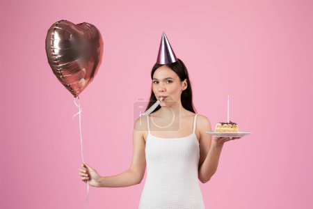 Foto de Mujer sonriente en sombrero de fiesta sosteniendo globo de corazón y una rebanada de pastel de cumpleaños, fiesta lista sobre fondo rosa - Imagen libre de derechos