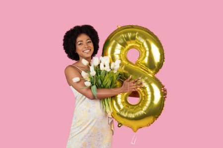 Téléchargez les photos : Une femme afro-américaine joyeuse tenant un ballon doré numéro 8 et des tulipes blanches fraîches célébrant un moment spécial - en image libre de droit