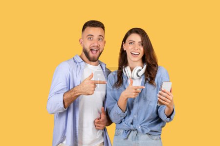 Téléchargez les photos : Jeune homme et femme pointant et regardant avec excitation un téléphone portable, sur un fond jaune - en image libre de droit