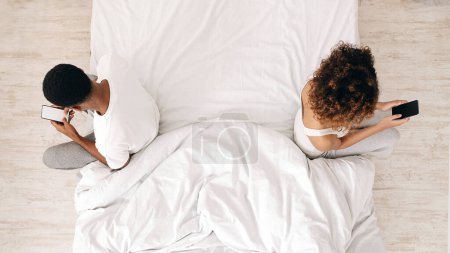 Photo for An overhead view of african american man and woman lying in bed, engrossed in their smartphones, depicting modern technology use in relationships - Royalty Free Image