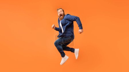 Photo for Joyful emotional man with red beard jumps in mid air and shouting in excitement, over orange studio background, smiling to camera. Concept of joy and good news celebration. Panorama, free space - Royalty Free Image