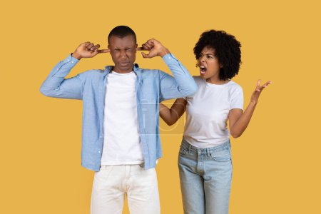 African American young couple are standing with one covering ears and the other shouting, suggesting a disagreement on a yellow background