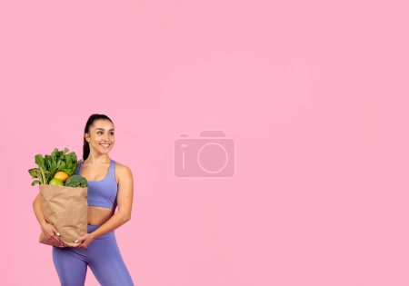 Téléchargez les photos : S'adapter jeune femme en vêtements de sport posant en toute confiance avec un sac d'épicerie, beaucoup d'espace de copie - en image libre de droit