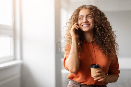 Foto de Una mujer alegre en una camisa naranja hablando en su teléfono móvil mientras sostiene una taza de café con iluminación natural, espacio para copiar - Imagen libre de derechos