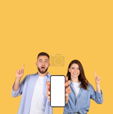 Téléchargez les photos : Homme et femme pointent avec enthousiasme vers un écran de smartphone vierge sur fond jaune - en image libre de droit