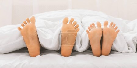 Foto de Pies desnudos de pareja en la cama. Pareja cariñosa acostada debajo de una manta blanca, panorama, espacio para copiar - Imagen libre de derechos