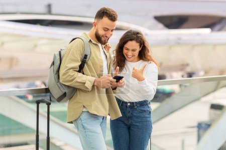 A smiling young couple is looking at a smartphone screen together, booking taxi online, standing at airport