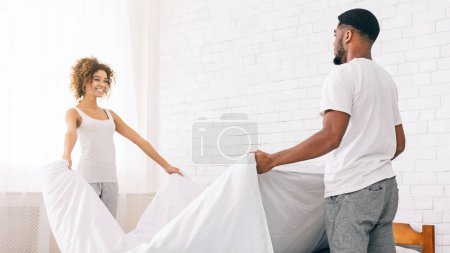 Ein fröhliches afrikanisch-amerikanisches Paar verbindet, während es das Bett zusammen in einem hellen, luftigen Schlafzimmer macht