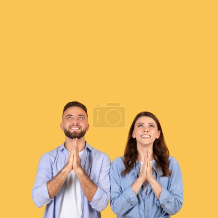 Ein fröhlicher Mann und eine fröhliche Frau stehen zusammen und blicken mit hoffnungsvoll zusammengefalteten Händen vor gelbem Hintergrund nach oben.