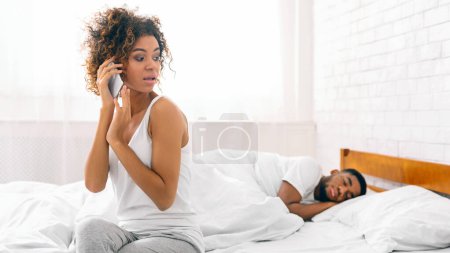 Mujer afroamericana habla por teléfono junto a su pareja dormida, retratando las complejidades de las relaciones modernas
