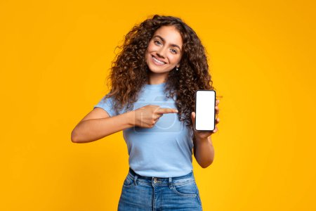Téléchargez les photos : Femme heureuse présentant un téléphone portable avec un écran blanc, exprimant la positivité sur un fond jaune - en image libre de droit
