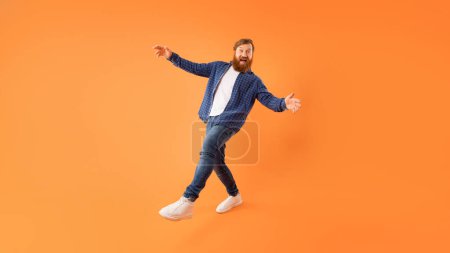Téléchargez les photos : Un mec aux cheveux roux positif avec la barbe en tenue décontractée qui se fait duper en posant sur un fond de studio orange, dansant et faisant des mouvements drôles, écartant ses bras large, plan complet, espace libre, panorama - en image libre de droit