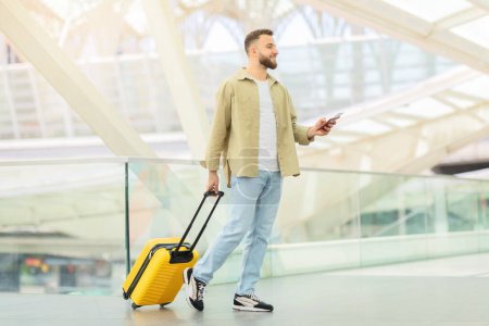 Photo for A fashionable man strolling with a bright yellow suitcase while using his phone at an airport - Royalty Free Image