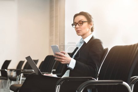 A focused businesswoman multitasks with a phone and laptop while waiting in a minimalistic and modern seating area