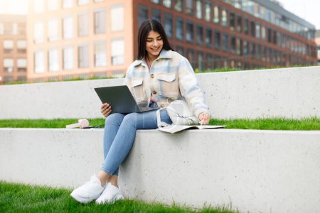 Cheerful young girl student sits on the curb with her laptop and notebook, studying in a relaxed urban setting