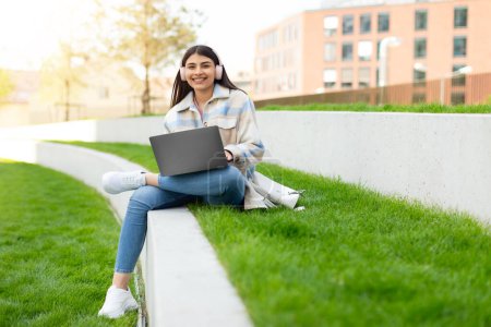 Foto de Una joven estudiante agradable sonríe ampliamente mientras está sentada con su computadora portátil en un banco urbano, con un libro a su lado - Imagen libre de derechos
