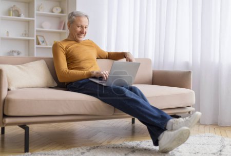 Téléchargez les photos : Un homme âgé s'assoit sur un canapé avec un ordinateur portable sur ses genoux, démontrant une concentration détendue dans un environnement familial - en image libre de droit