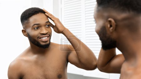 Ein zufriedener afroamerikanischer Mann bewundert sein Spiegelbild im Badezimmerspiegel und zeigt Selbstbewusstsein und Positivität