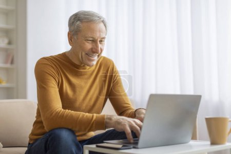 Téléchargez les photos : Homme d'affaires plus âgé avec un sourire rayonnant travaille sur un ordinateur portable dans un environnement confortable salon à la maison - en image libre de droit