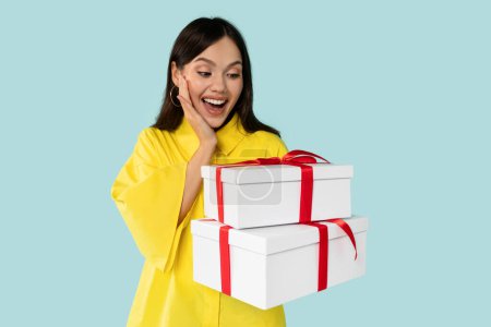 Emotional überraschte junge brünette Frau steht und hält weiße Geschenkboxen mit roten Schleifen, trägt gelbes Hemd vor blauem Hintergrund, Konzept für Geburtstagsfeier