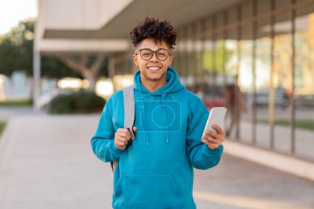 Ein fröhlicher brasilianischer Student mit stylischer Brille und Kopfhörer um den Hals hält ein Smartphone, urbaner Hintergrund