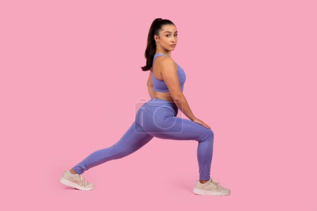 Fit young woman in athletic wear demonstrating a lunge pose against a pink background, showcasing exercise form
