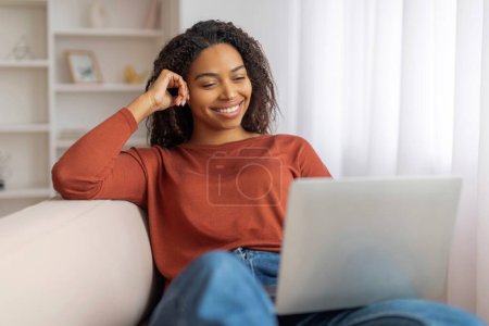 Téléchargez les photos : Femme souriante noire assise confortablement sur un canapé tout en utilisant un ordinateur portable, heureuse femme afro-américaine regardant des films en ligne ou naviguant sur Internet tout en se reposant sur le canapé - en image libre de droit
