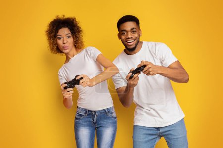 Téléchargez les photos : Joyeux couple afro-américain jouissant de jeux vidéo, jouant avec des joysticks sur fond de studio orange - en image libre de droit