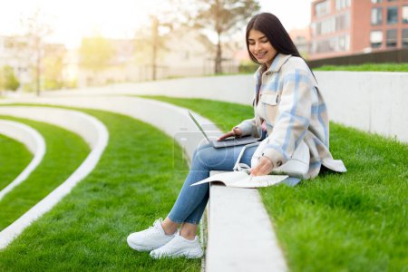 Eine junge Frau strahlt vor Freude, während sie einen Laptop benutzt und ein Smartphone in der Hand hält, bequem im Freien sitzend