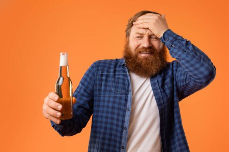 Redhaired bearded man with unhappy expression holding beer bottle and touching head, suffering from headache during hangover, standing over orange studio backdrop. Negative effects of alcohol