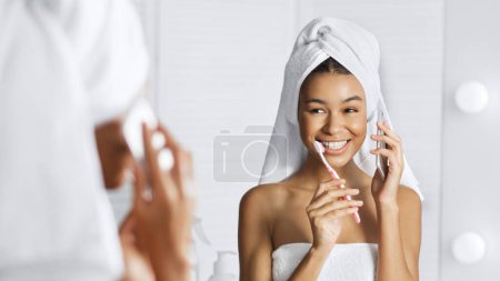 Photo for Cute afro-american girl talking by phone and cleaning teeth simultaneously in front of mirror in bathroom. Girls morning routine concept. - Royalty Free Image