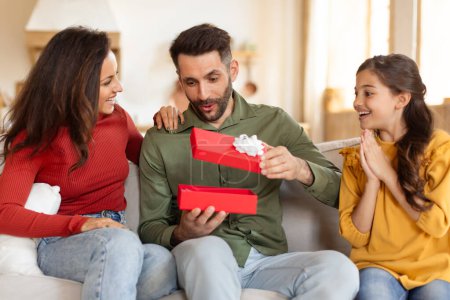 Foto de Emocionado padre desenvainando un regalo rojo con su familia, capturando un momento de alegría y sorpresa en el sofá - Imagen libre de derechos