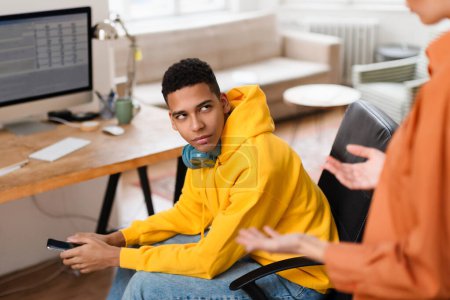 Foto de Un joven con una sudadera amarilla sostiene un teléfono inteligente mientras interactúa con una persona invisible en un entorno de oficina en casa - Imagen libre de derechos