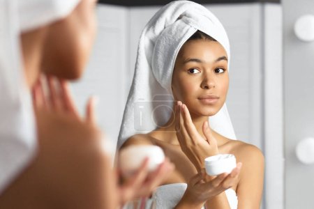 Téléchargez les photos : Afro-américaine fille tenant visage crème produit hydratant peau après douche debout dans la salle de bain. Concept de soins de la peau. Panorama - en image libre de droit