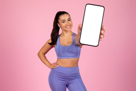 Téléchargez les photos : Une femme sportive en tenue de fitness souriante et présentant un écran de smartphone vierge, idéal pour les promotions d'applications - en image libre de droit