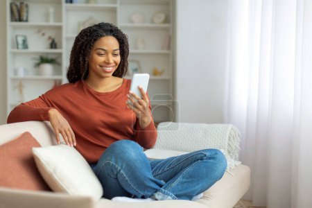 Photo for Black woman seated on a couch indoors, african american female engrossed in using a cell phone, messaging online or browsing app - Royalty Free Image