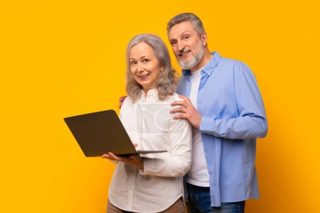 Téléchargez les photos : Un mari et une femme âgés souriants debout avec un ordinateur, engagés dans l'apprentissage en ligne et la vidéo appelant sur leur ordinateur portable, faisant défiler les médias sociaux et surfant ensemble sur un fond de studio jaune - en image libre de droit