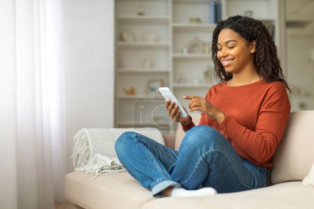 Téléchargez les photos : Joyeuse femme noire assise avec désinvolture sur un canapé, femme afro-américaine engagée avec son smartphone, souriant tout en envoyant des messages sur son téléphone portable - en image libre de droit