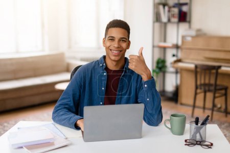 Foto de Joven adolescente negro alegre que estudia con el ordenador portátil en el ajuste casero, dando pulgares hacia arriba a la cámara, incorporando positividad y éxito en la educación - Imagen libre de derechos