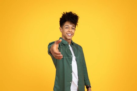 An exuberant young african american man extends a hand in greeting against a simple yellow backdrop, inviting interaction