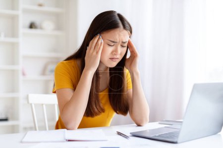 Foto de Joven mujer asiática sintiendo estrés y dolor de cabeza mientras trabaja en el ordenador portátil en casa oficina, frotando sus sienes - Imagen libre de derechos