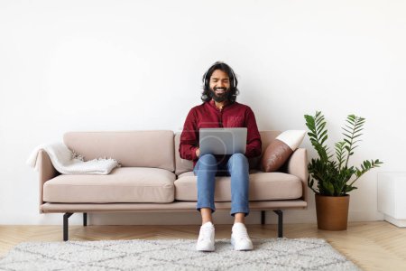 Téléchargez les photos : Heureux Indien assis sur un canapé avec un ordinateur portable sur ses genoux, exsudant joie et satisfaction dans un salon bien aménagé - en image libre de droit