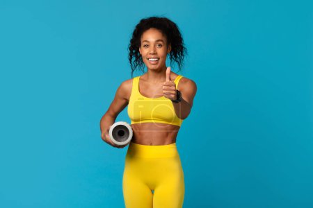 Fit African American woman in yellow active wear giving thumbs up and holding a yoga mat on a blue background