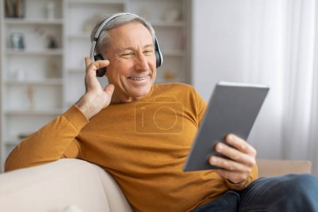 Téléchargez les photos : Homme âgé souriant dans un cadre confortable à la maison avec écouteurs, profiter de la technologie et de la musique sur une tablette - en image libre de droit