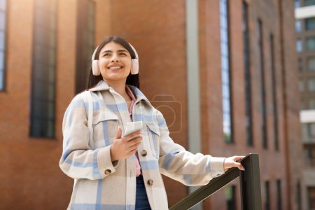 Téléchargez les photos : Une jeune fille souriante aime la musique à travers des écouteurs, tenant un smartphone avec des bâtiments en arrière-plan - en image libre de droit