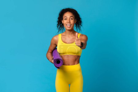 Téléchargez les photos : Femme afro-américaine souriante tenant un tapis de yoga violet donnant un pouce en vêtements de sport jaunes sur un fond bleu - en image libre de droit